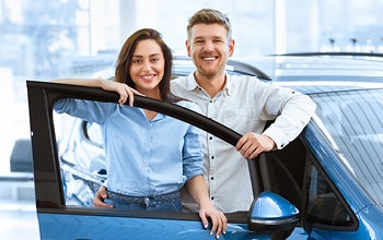 A man and woman stand together in front of a parked car, smiling and engaged in conversation.